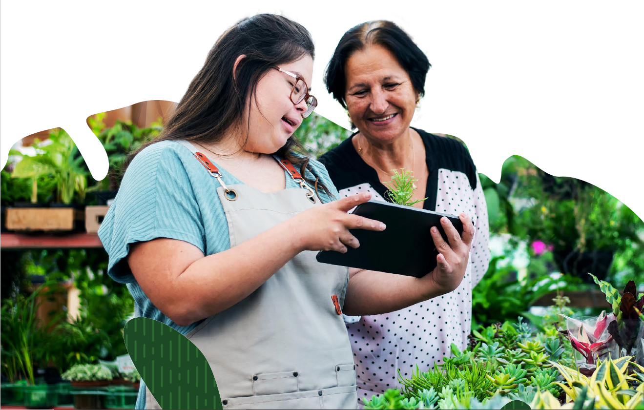 Two women in aprons work together on an electronic tablet in a greenhouse