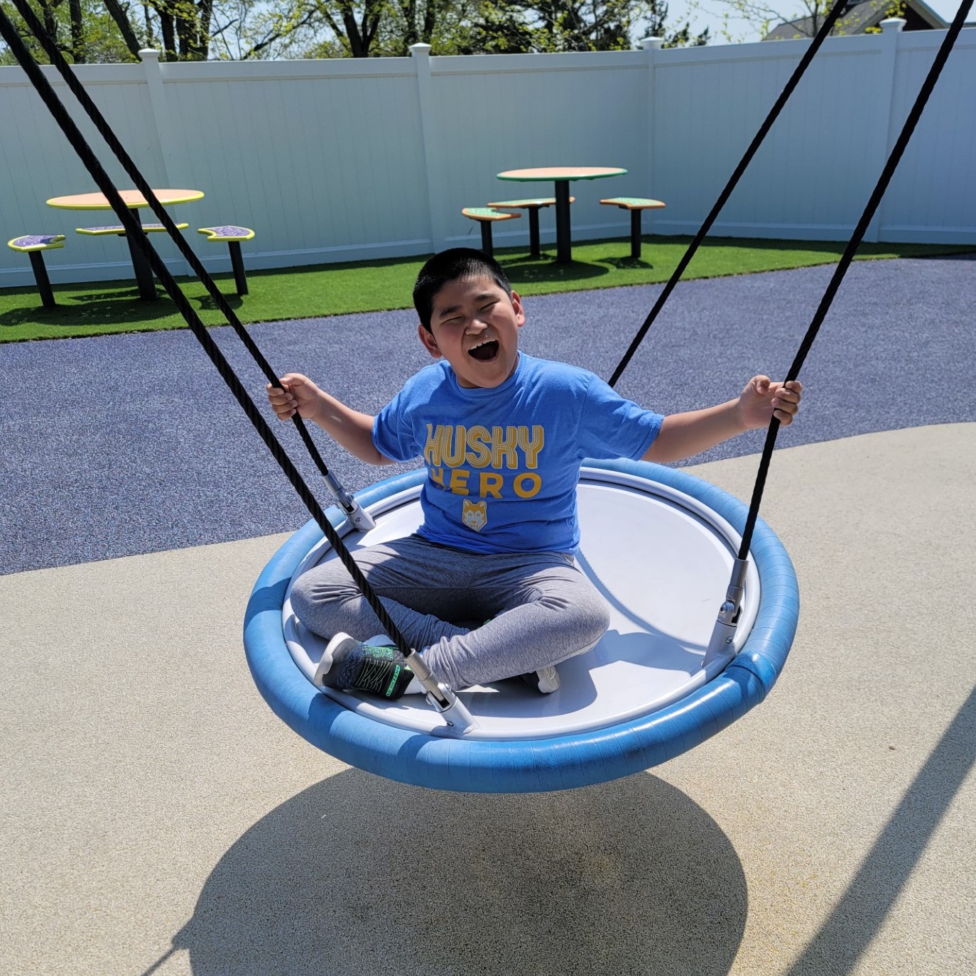 A student smiles widely as he sits on an adaptive swing set at The Guild School playground on a sunny day.