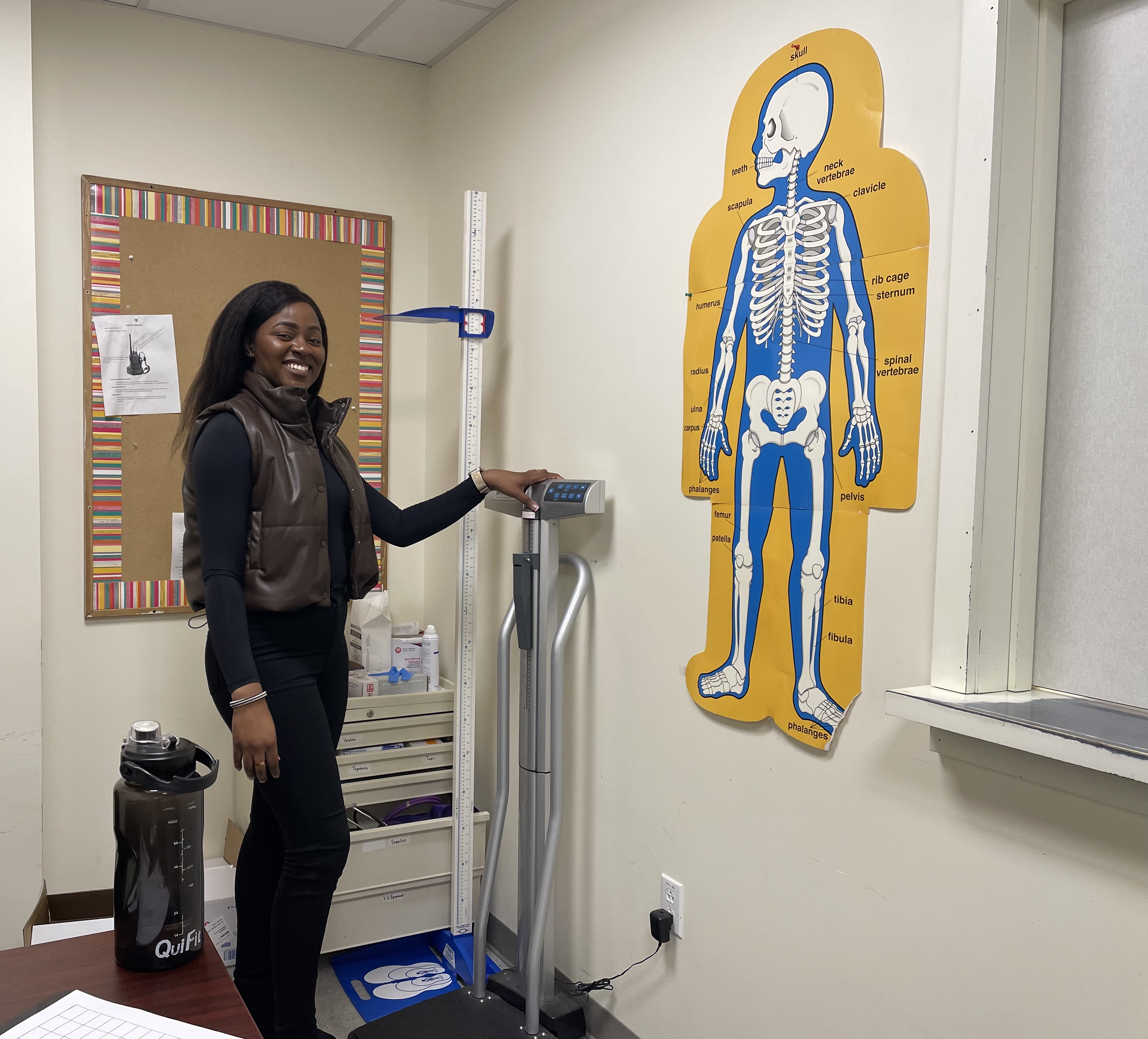 Sandra Kalambayi the School Nurse stands in her office with medical devices and a poster of the human skeletal system.