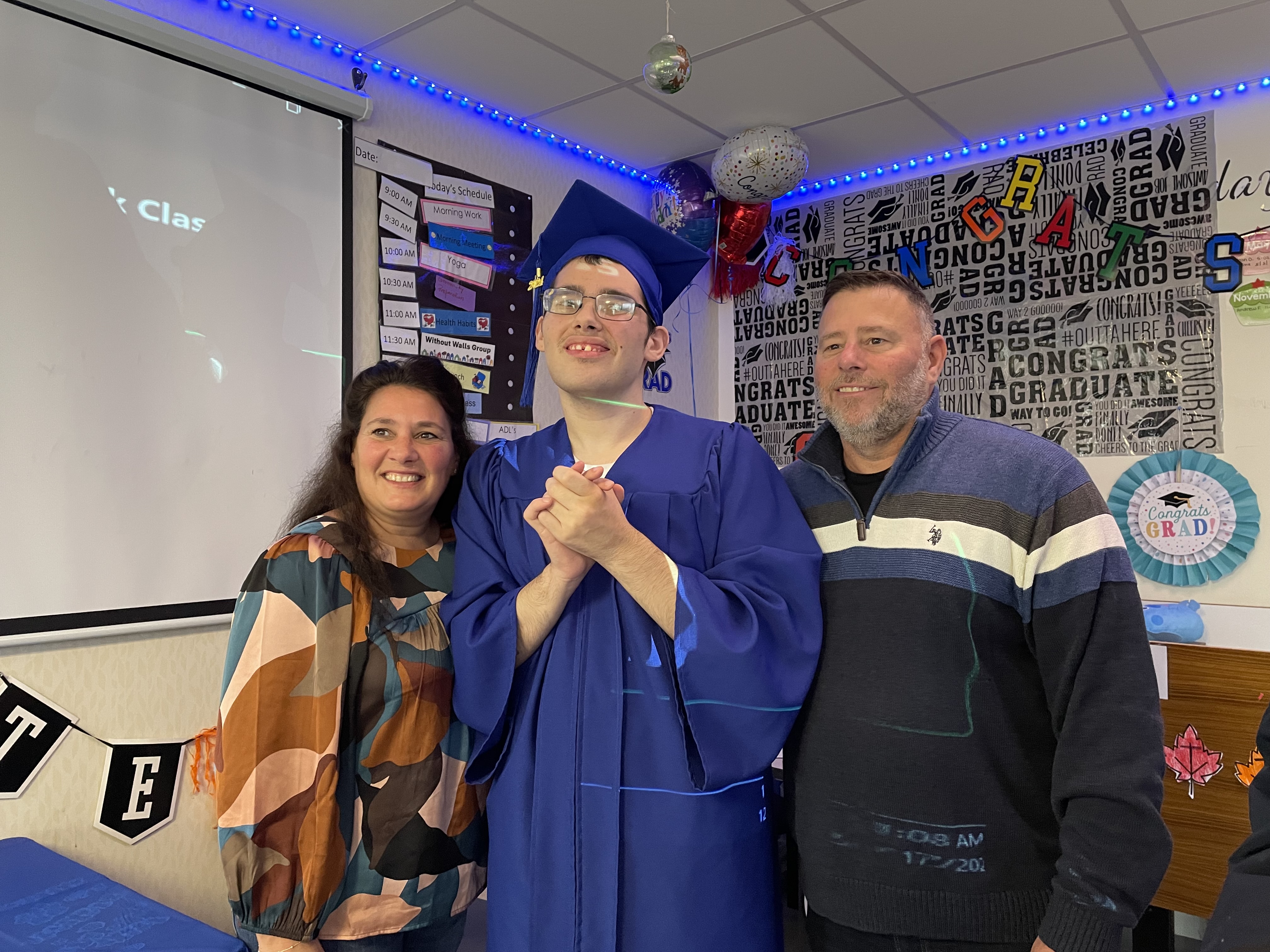 A graduate stands happily with hands clasped in a blue graduation gown and cap with his mom and dad on either side of him.