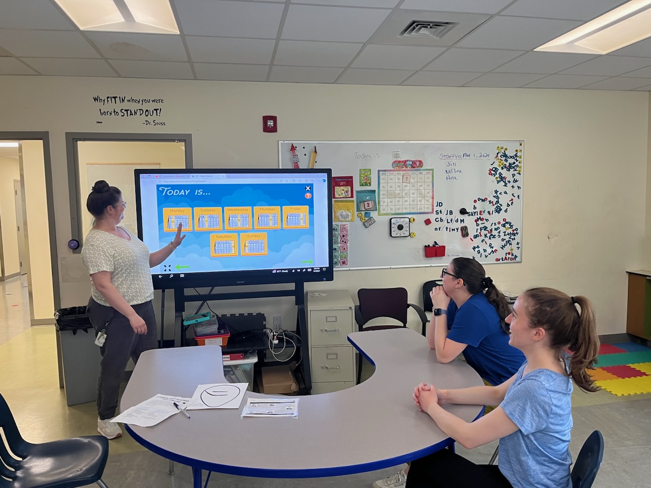 A teacher stands in the front of a classroom and points at a screen with a visual schedule while two people sit at a curved table looking on.