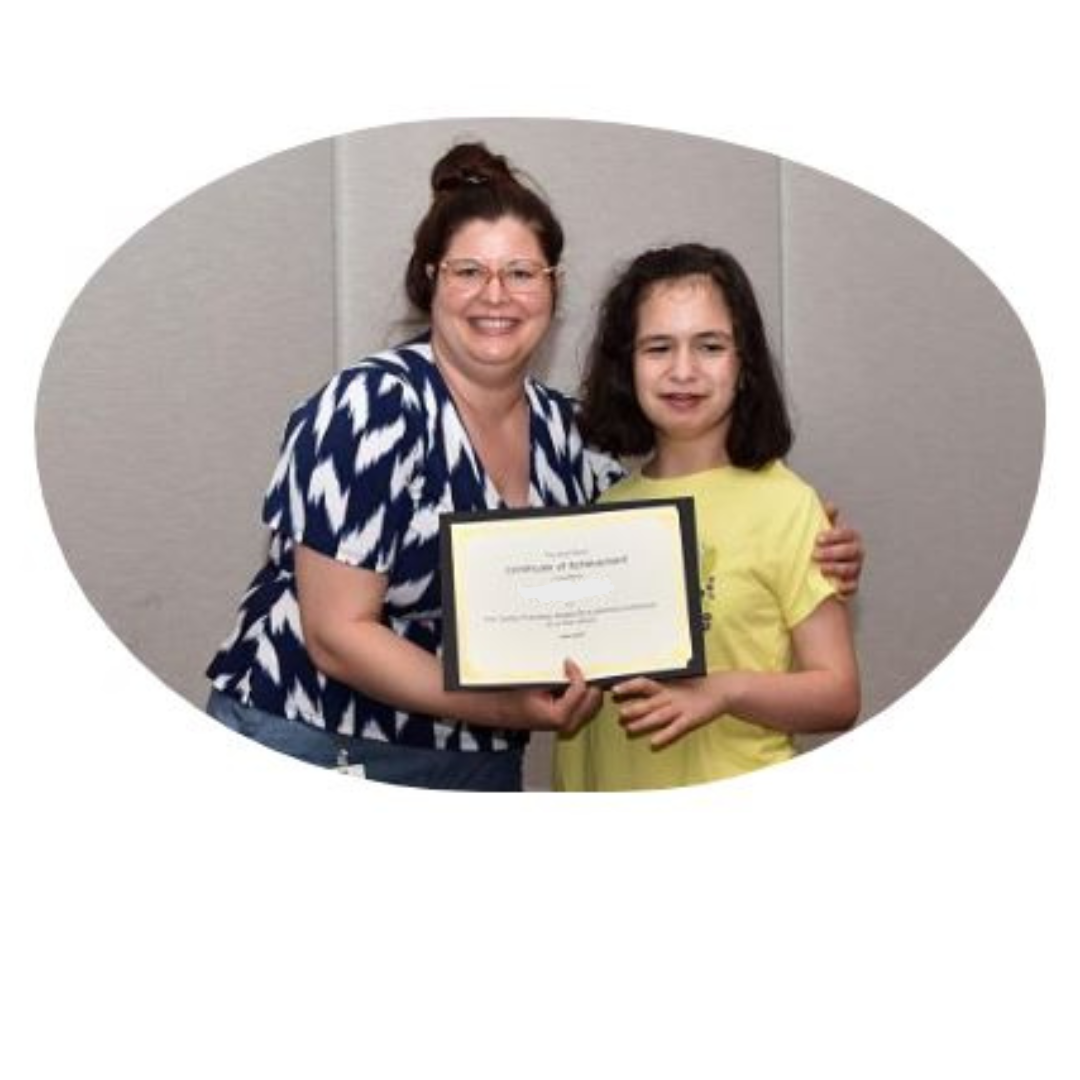 A staff member in a blue and white striped shirt helps a young woman in a bright yellow t-shirt hold up an award certificate