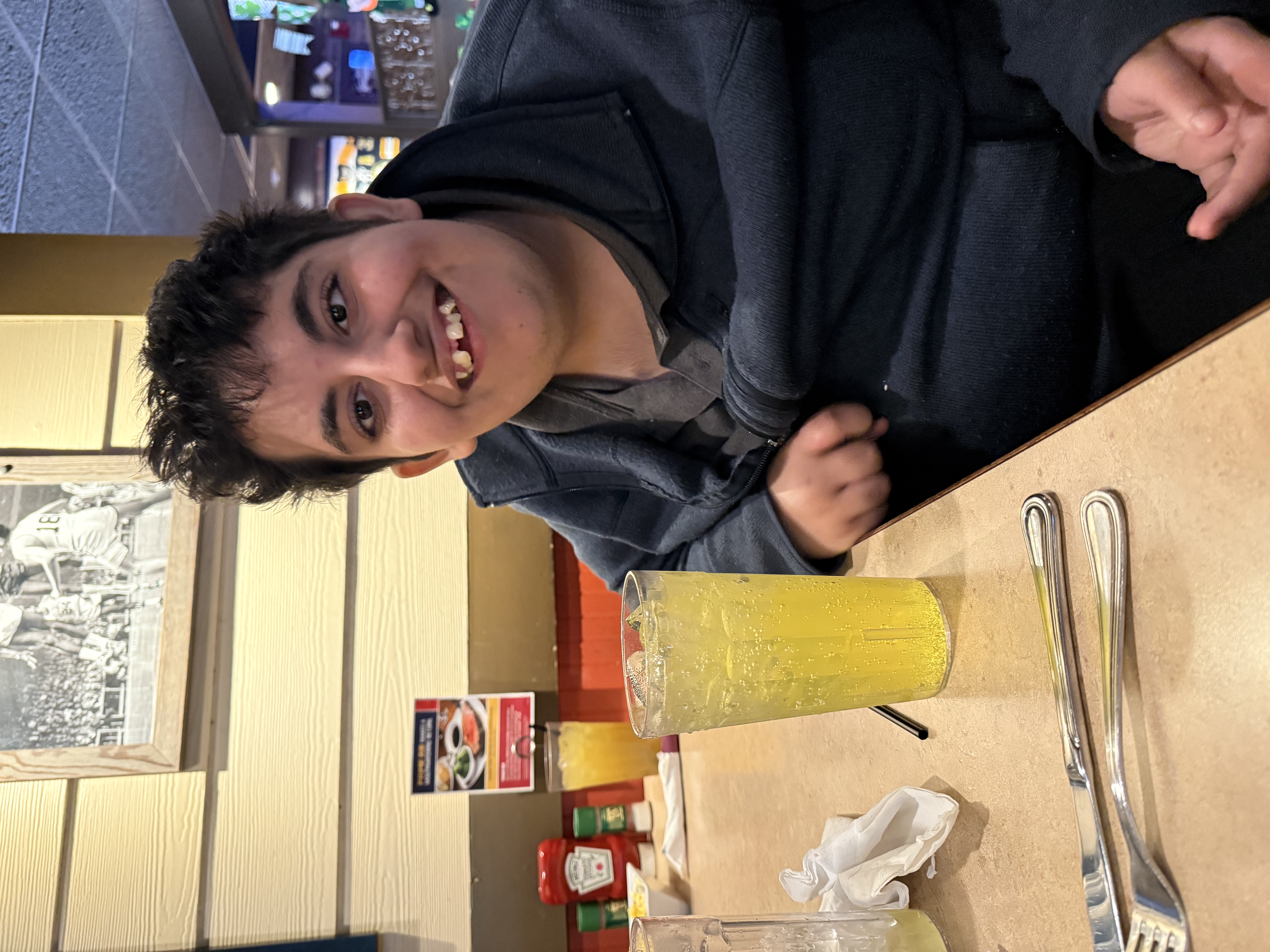 A student smiles into the camera while sitting with a large drink order at a restaurant table.