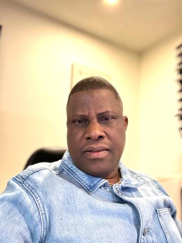 Olugesun sits in an office chair in front of a beige wall, looking directly into the camera with a neutral expression.