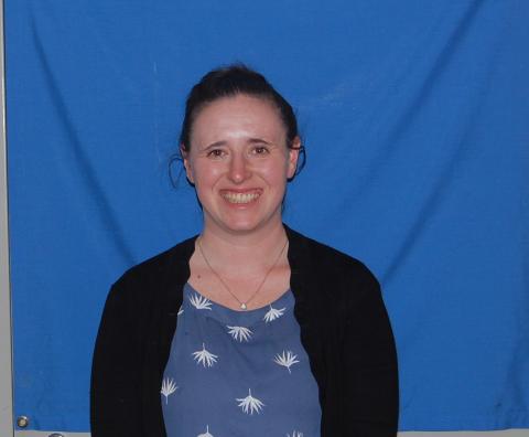 A photo of Nicole Picone smiling at the camera. She is wearing a blue shirt with white print and a black cardigan, and she stands in front of a blue backdrop.