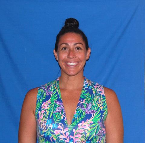 Christina Preville smiles into the camera. She is wearing a bright sleeveless shirt with teal, green, and pink flowers and leaves. She stands in front of an all-blue backdrop.