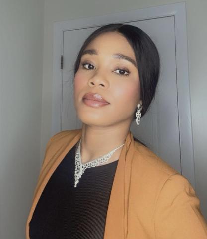 Mercy Jolo looks into the camera with a neutral expression. She wears her hair up and a tan cardigan over a simple black shirt. Behind her is a white wall and white door.