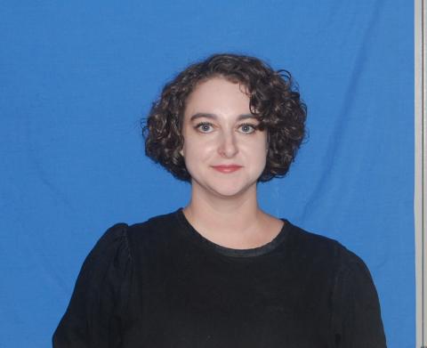 Courtney Dumais smiles slightly into the camera. She is wearing an all black shirt and stands in front of a blue backdrop.