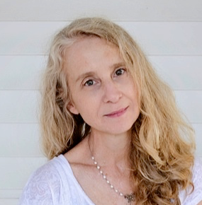 Mary Barnes looks directly into the camera with a neutral expression. She is wearing a white shirt and silver necklace in front of a plain white wall.