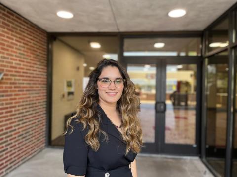 Naseema Amin stands in front of a brick building with large glass doors and windows. She has long, wavy hair and wears thin frame glasses and a black top.