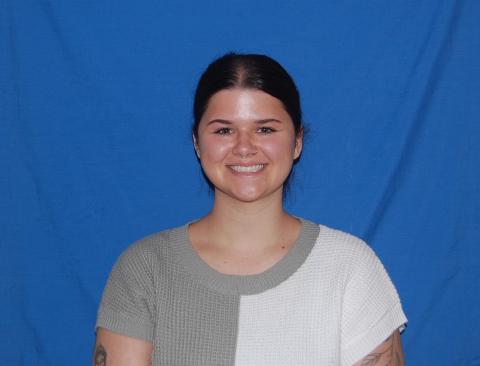 Emily Shepard stands in front of a plain blue background. She is smiling with her hair in a ponytail and wearing a white and gray t-shirt.