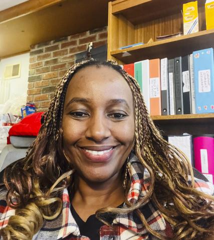 Maureen Kemigisha smiles casually into the camera. She has long curly, partially-braided hair and sits in front of a bookshelf.