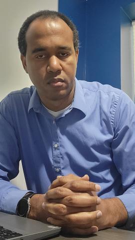 Nansen Alexandre looks into the camera with a neutral expression and folded hands. He is wearing a light blue shirt and sits in front of a darker blue wall.